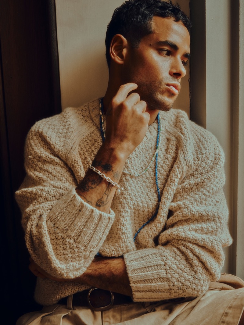 Washington wears a cream textured cable-knit crewneck sweater with khaki pleated shorts, a brown leather belt, a blue beaded necklace, and a silver chain bracelet for Banana Republic's Archive Reissue collection