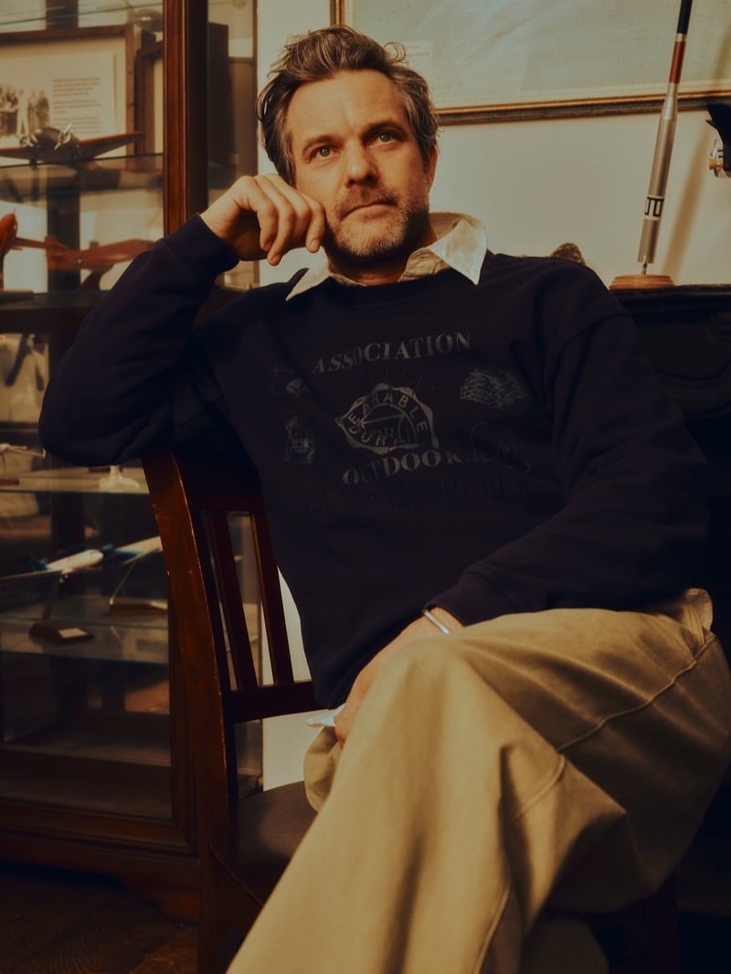 Jackson sits in a navy Outdoor Association graphic crewneck sweatshirt over a white shirt with wide-leg khaki trousers, beside a glass case of model airplanes for Banana Republic's Archive Reissue collection