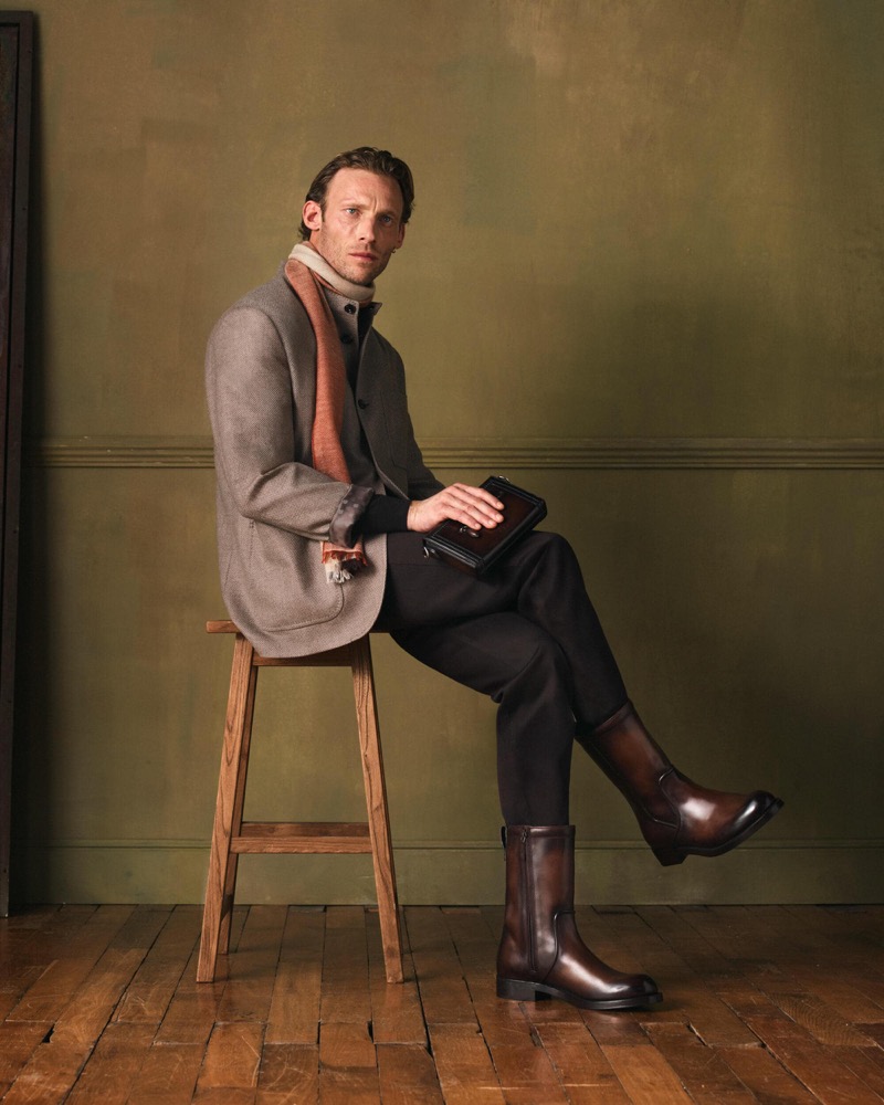 Arnaud sits on a wooden stool in a grey overcoat with a peach scarf, black trousers, and brown Venezia calfskin mid-calf boots