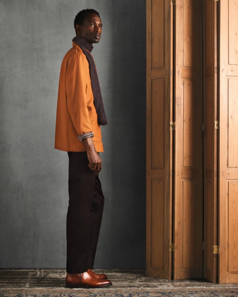 Moussa stands in profile wearing an orange jacket with a dark scarf, black trousers, and cognac leather shoes