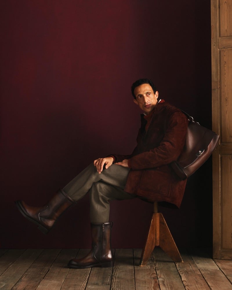 Naoki sits on a stool in a dark brown suede coat over olive trousers with brown mid-calf boots and a leather bag