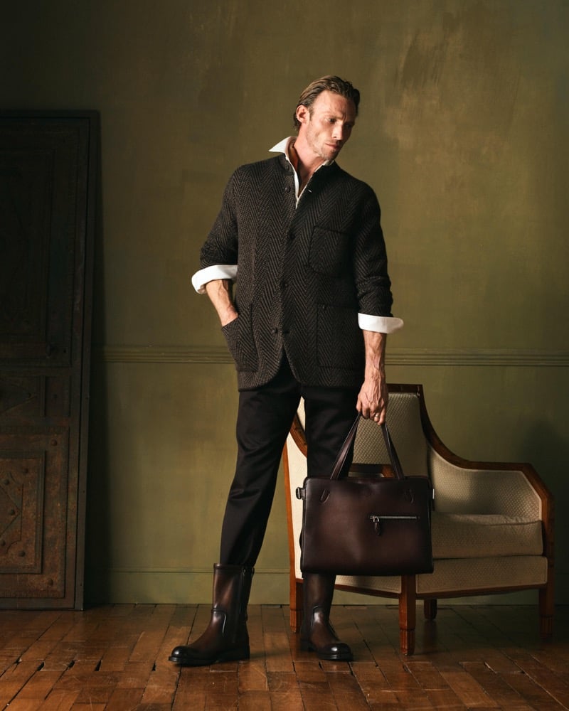 Arnaud stands in a dark herringbone jacket over a white shirt with black trousers, brown mid-calf boots, and a dark leather bag
