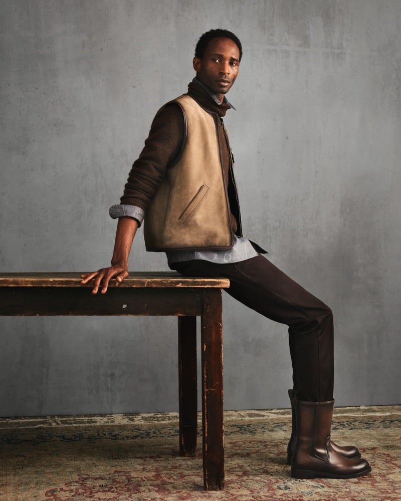 Moussa leans against a wooden table in a tan-and-brown shearling vest over a shirt with dark trousers and brown boots