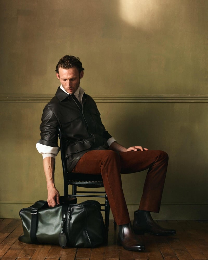 Arnaud sits on a chair in a dark leather jacket over a white shirt with rust corduroy trousers, black Chelsea boots, and a green leather bag