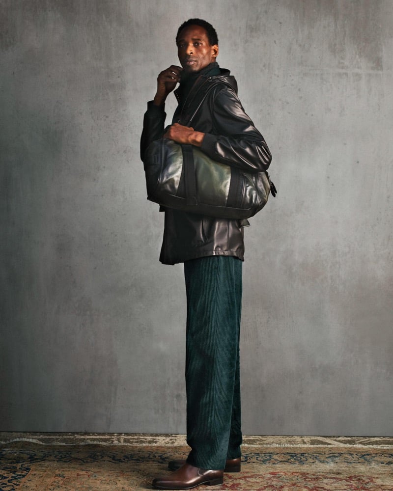 Moussa wears a black leather hooded jacket with a dark leather bag over teal corduroy trousers and brown leather shoes