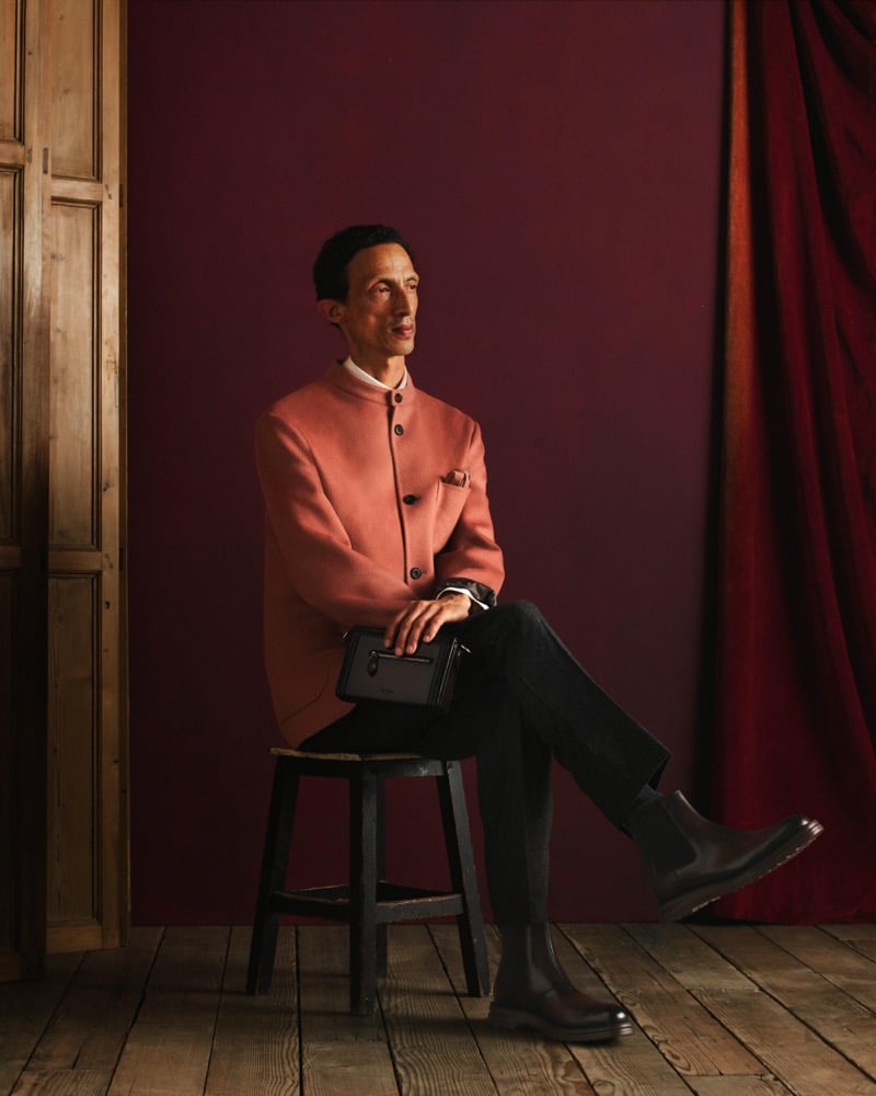 Naoki sits on a stool in a salmon-pink Nehru-collar jacket over a white shirt with dark trousers, black boots, and a small leather case
