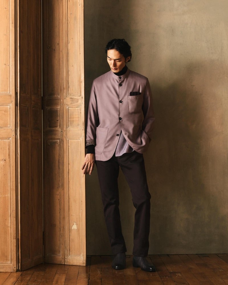 Naoki stands by a wooden door in a lavender band-collar jacket over a dark shirt with dark trousers and black shoes