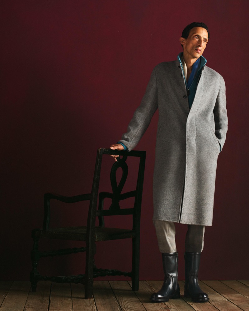 Naoki stands beside a chair in a grey overcoat over a teal jacket with beige trousers and black mid-calf boots