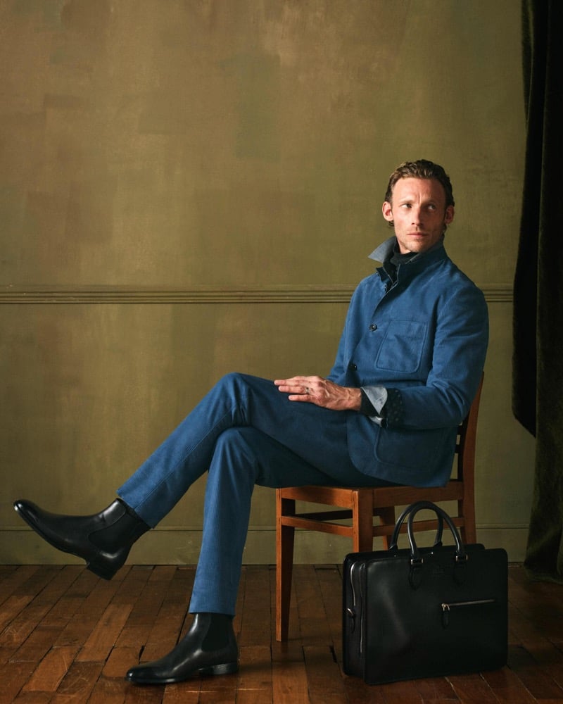 Arnaud sits on a chair in a blue cotton suit with a grey collar shirt, black Chelsea boots, and a black leather briefcase