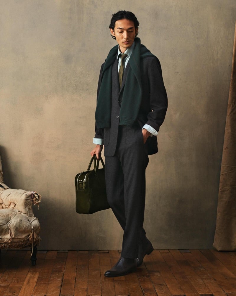Naoki wears a dark jacket with a green scarf and tie over a white shirt with charcoal trousers, black shoes, and a green leather bag