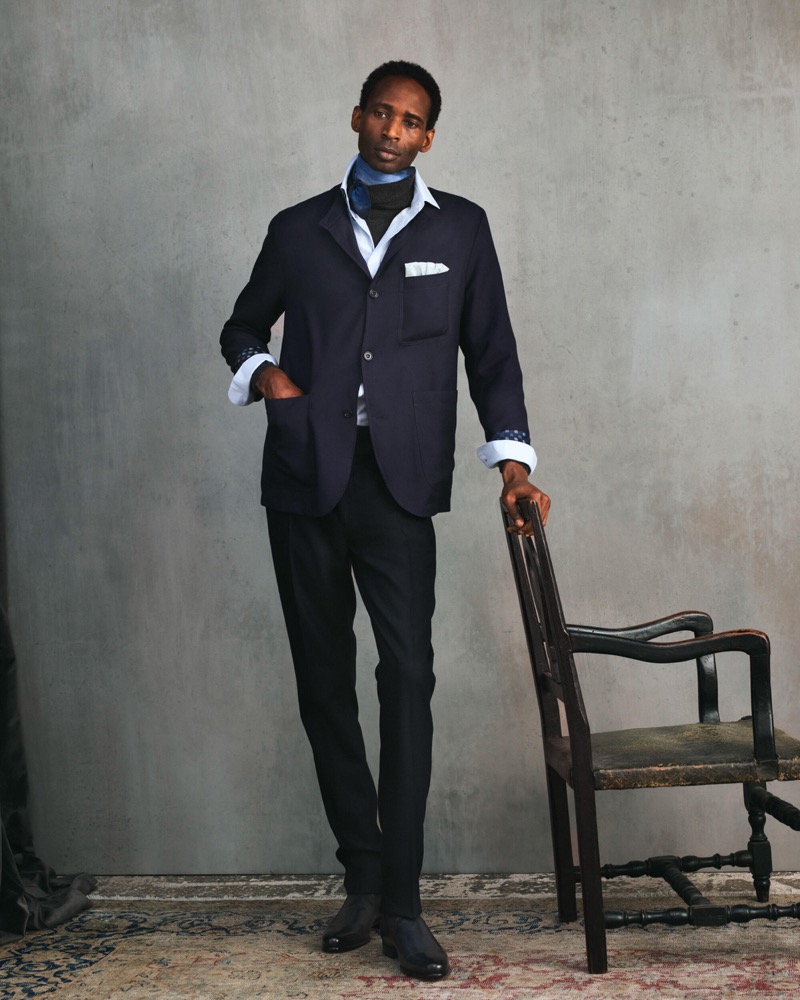 Moussa stands beside a chair in a navy blazer with a blue scarf and pocket square over a light shirt with black trousers and dark boots