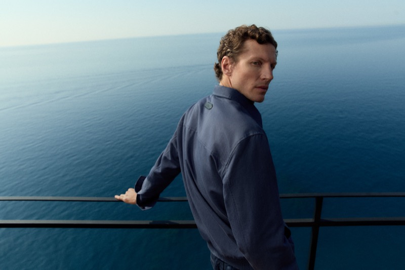 Alex looks over his shoulder wearing a navy camp collar shirt with the Amalfi coast sea behind him