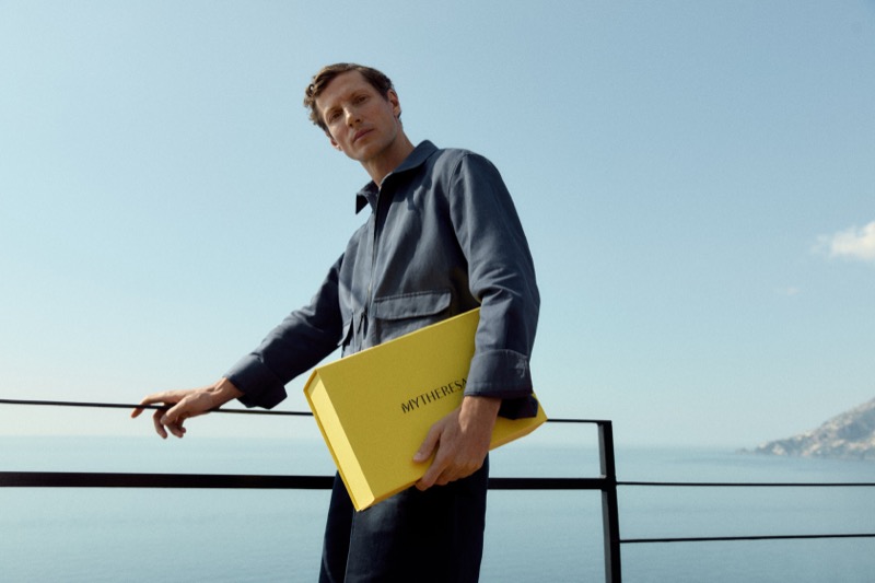 Alex holds a yellow Mytheresa box while wearing a navy camp collar shirt on a coastal terrace