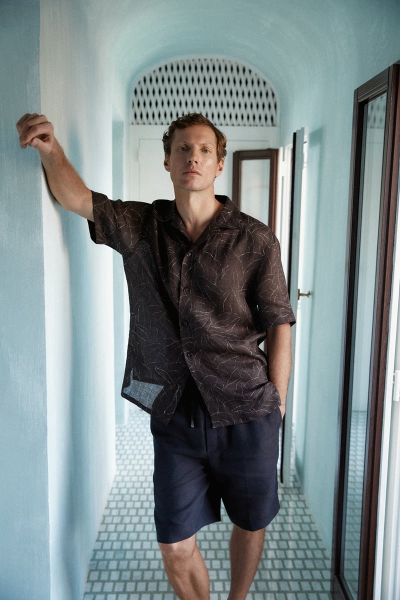 Alex leans against a white hallway wall wearing a brown printed camp collar shirt and navy shorts