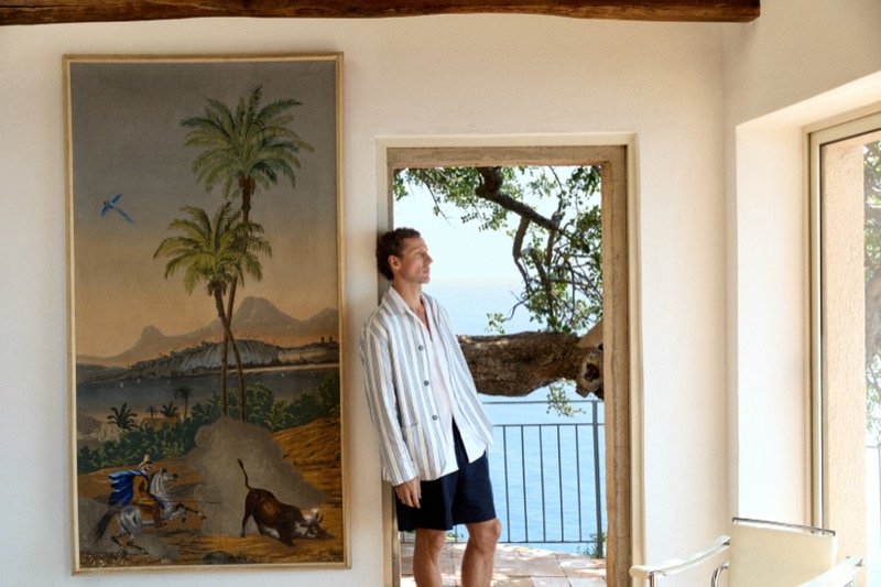 Alex stands beside a tropical painting wearing a striped overshirt and navy shorts