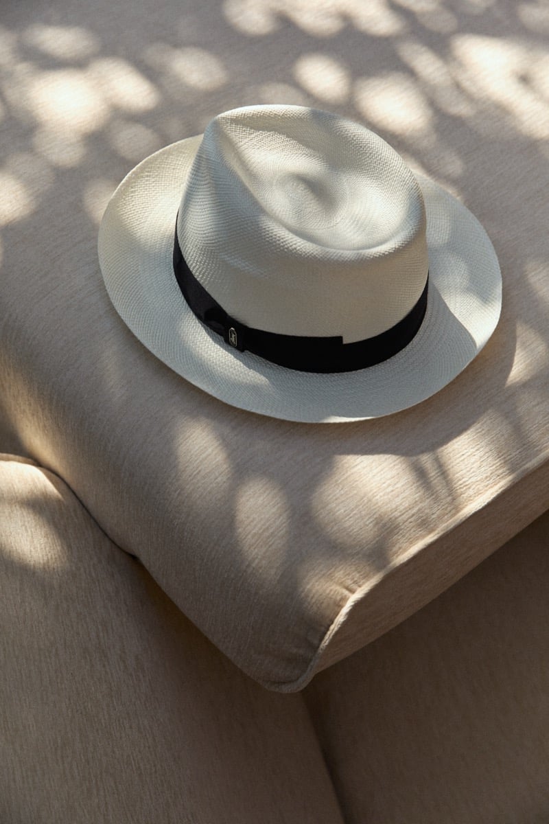 A white Panama hat with a black grosgrain band rests on a linen cushion in dappled afternoon light
