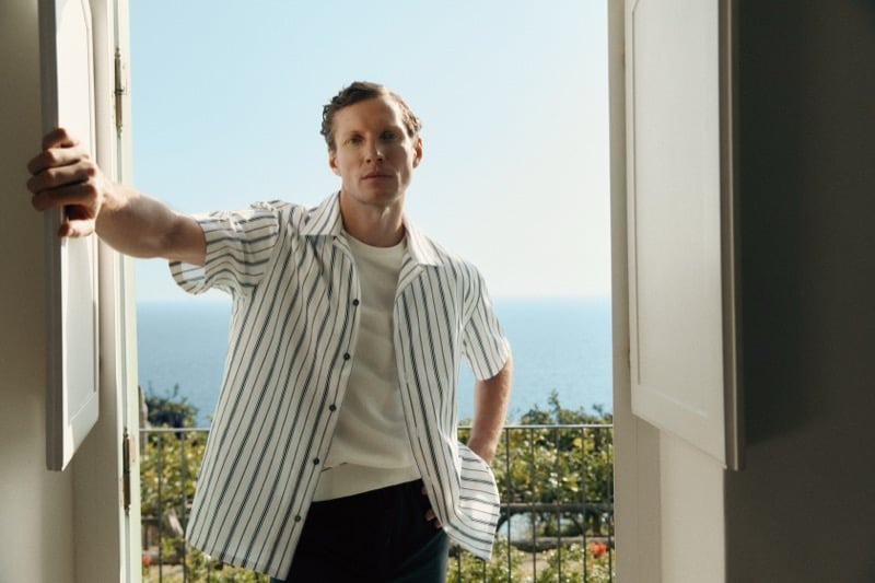 Alex stands in a doorway wearing a striped linen overshirt over a white T-shirt with the sea visible behind him