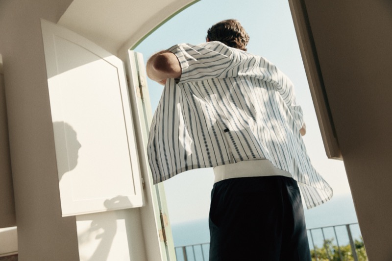 Alex steps through a doorway from behind wearing a striped overshirt with sunlight casting shadows