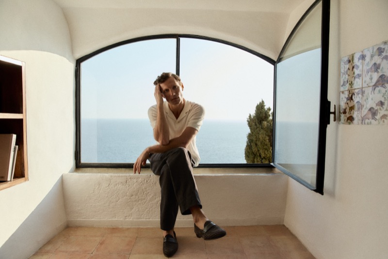 Alex sits in an arched window overlooking the sea wearing a white shirt and dark trousers with woven leather loafers
