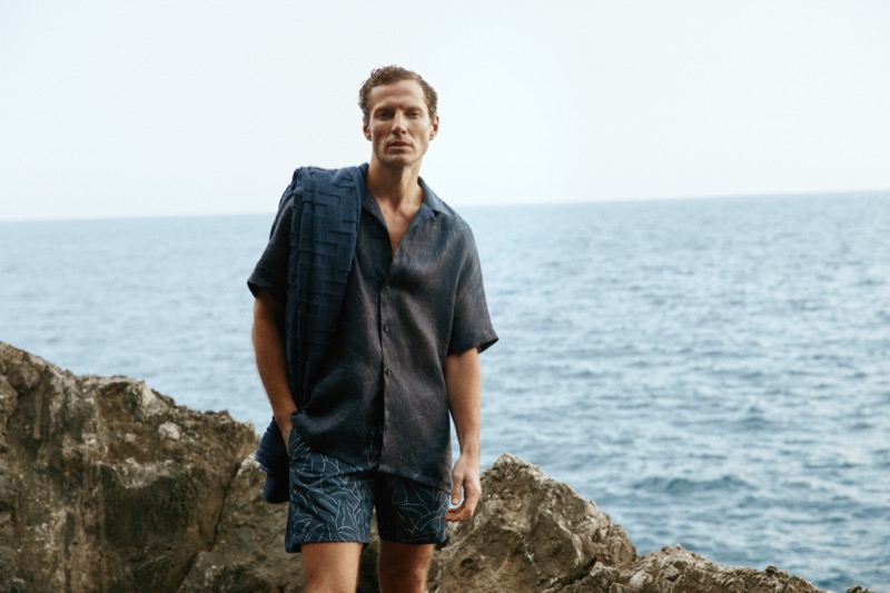 Alex stands on coastal rocks wearing a navy camp collar shirt, patterned swim shorts, and a towel over his shoulder
