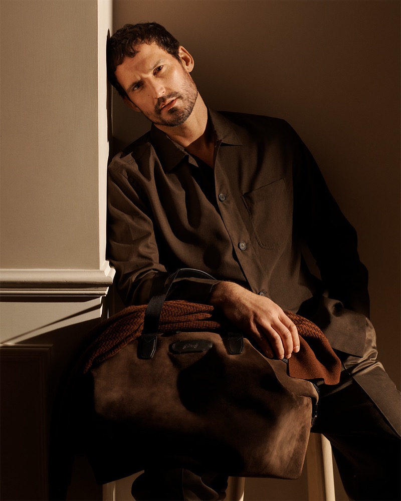 Sam wears a Brioni dark olive overshirt and holds a dark suede travel bag with a woven leather strap