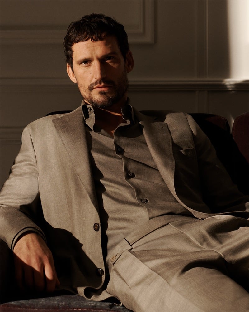 Sam wears a Brioni taupe linen suit with a matching shirt, seated in warm amber interior light