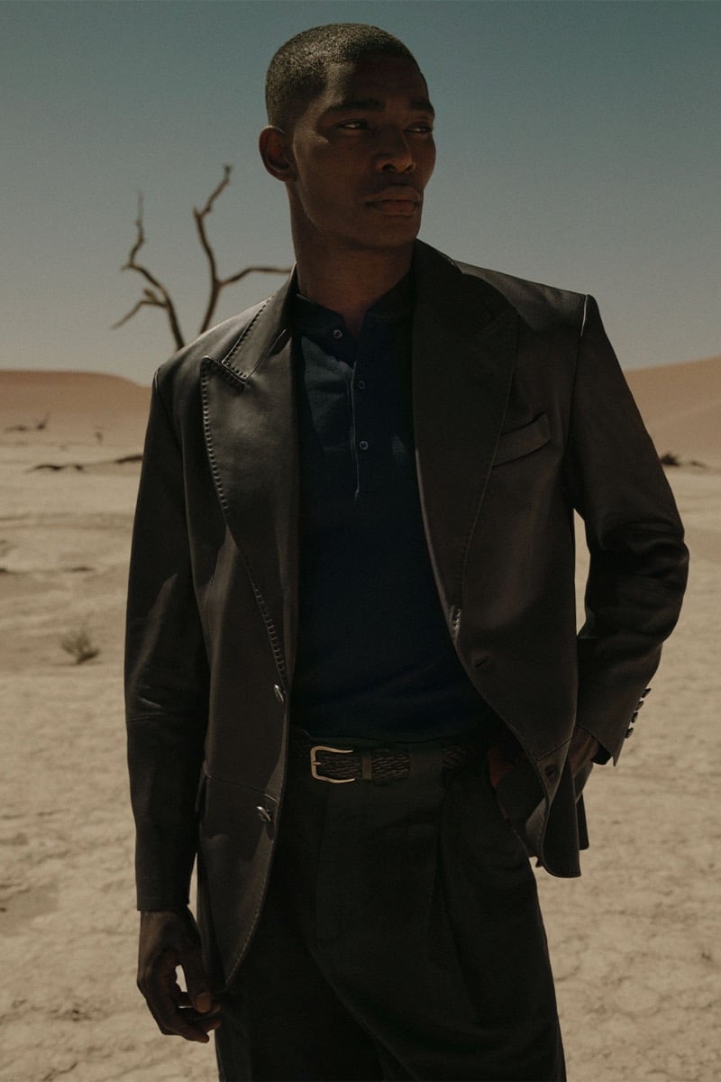 Niyo wears a dark leather blazer over a navy henley with dark trousers and a woven belt