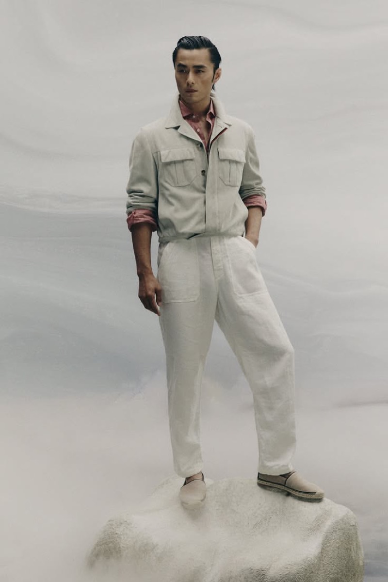 Zhao wears an off-white suede jacket over apricot-colored linen with white trousers standing on a rock in mist