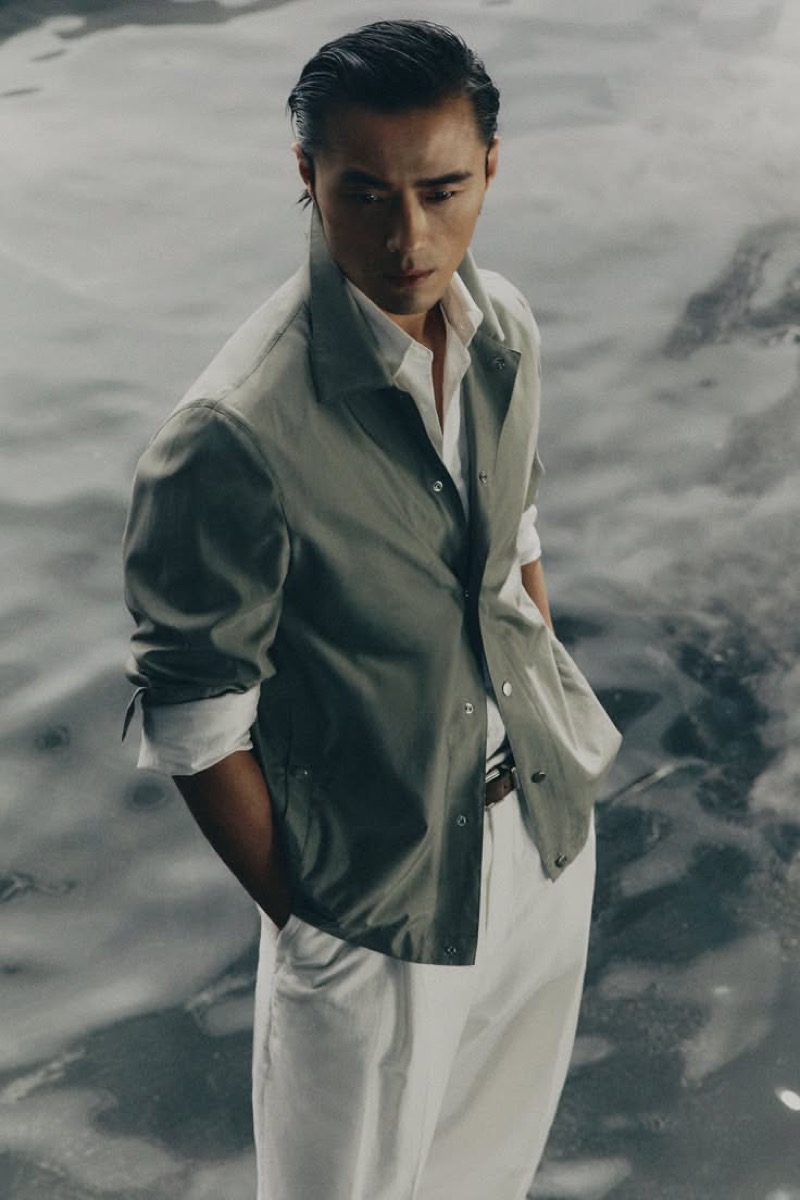 Zhao wears a silk khaki jacket with a white shirt and trousers against a water backdrop