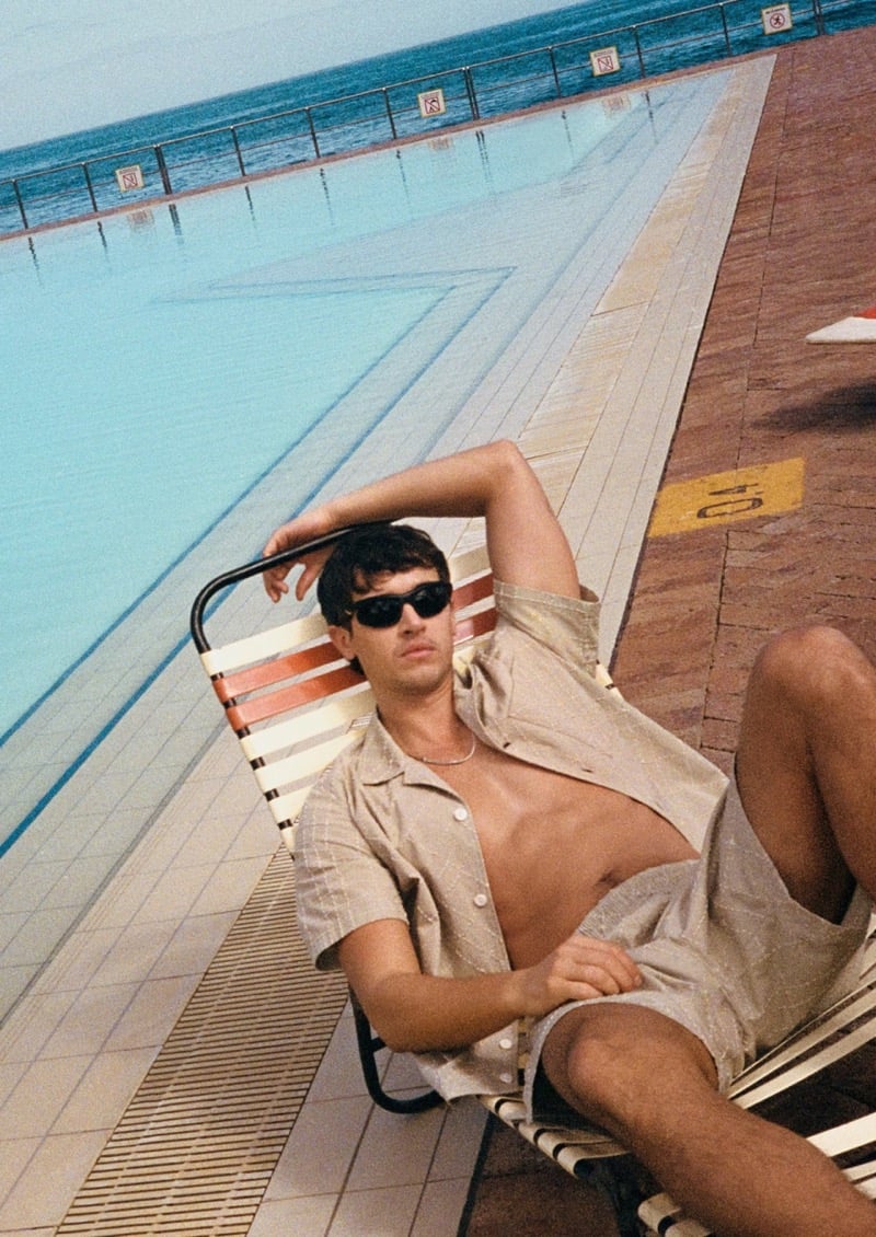 Blyth reclines on a lounge chair poolside in an open short-sleeve shirt and shorts, wearing dark sunglasses and a chain necklace, for the Burberry high summer 2026 campaign
