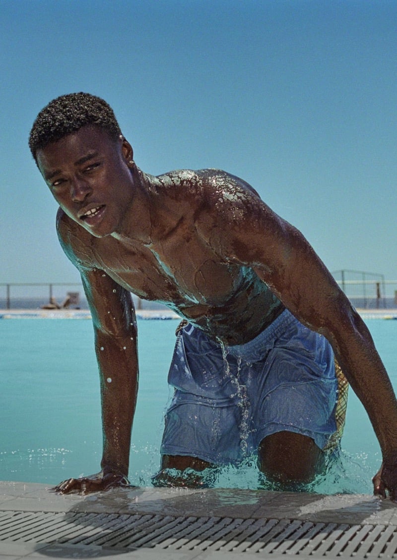 N'Doye, shirtless and wet, climbing out of a lido pool in blue Burberry check swim shorts under a bright blue sky for the high summer 2026 campaign