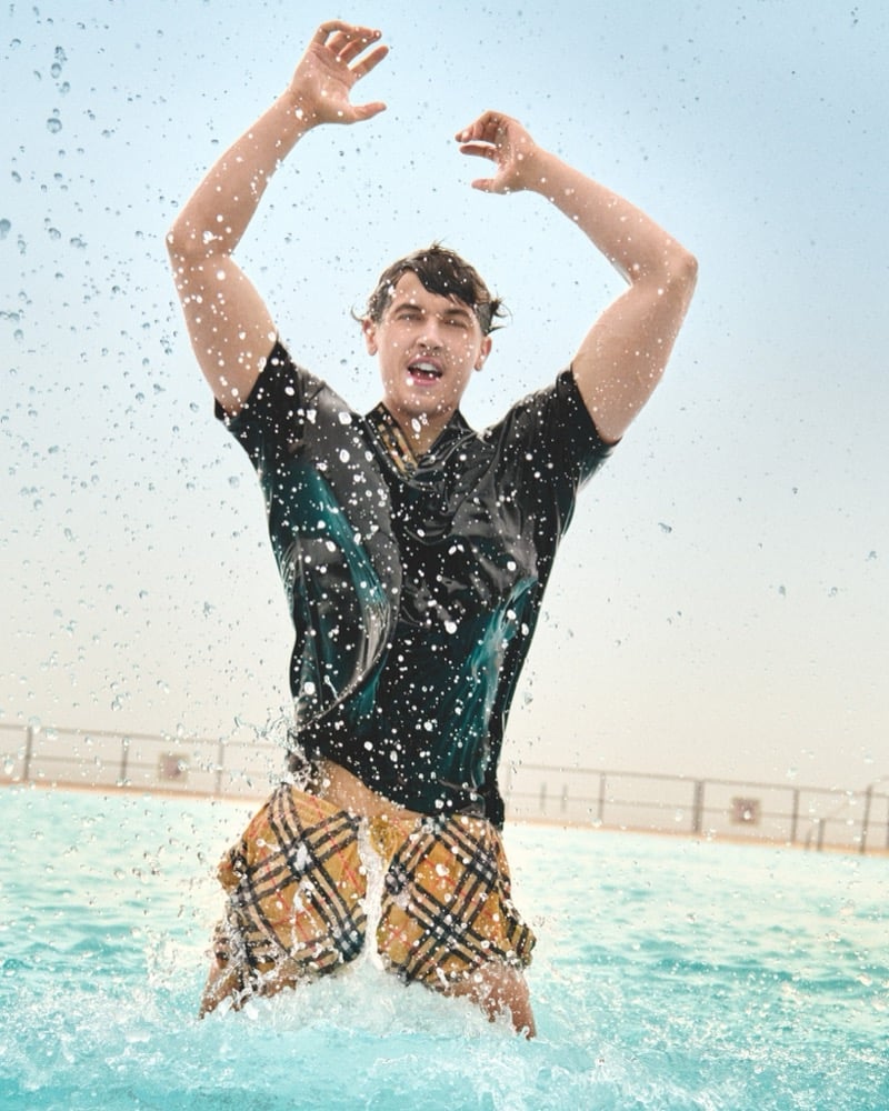 Blyth stands in a pool with arms raised and water splashing around him in a dark polo and Burberry check swim shorts for the high summer 2026 campaign
