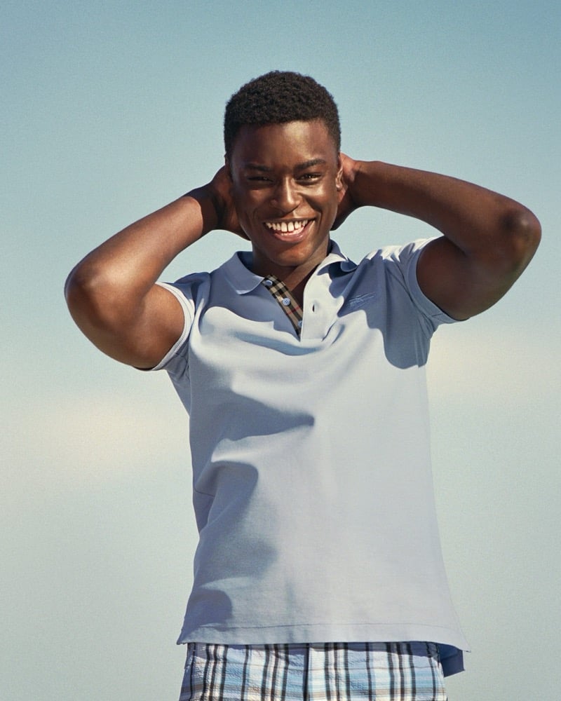 N'Doye laughs with hands behind his neck in a cornflower blue Burberry polo with check collar detail and check shorts against a pale blue sky for the high summer 2026 campaign