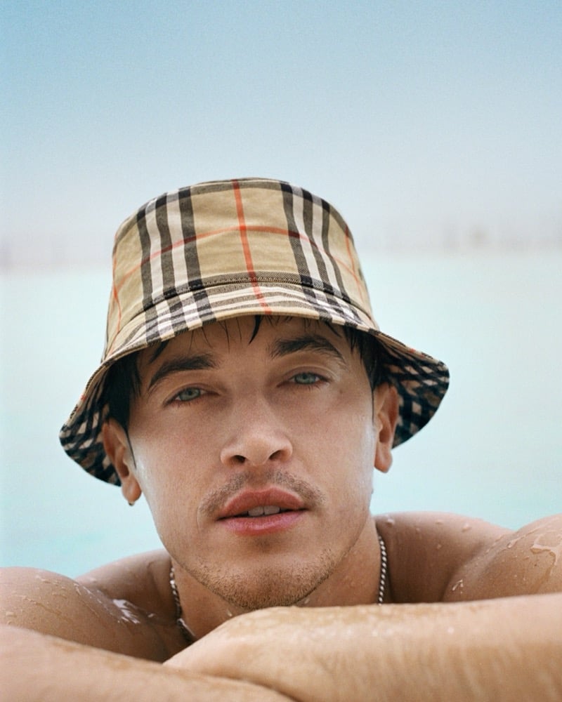 Tom Blyth rests at the edge of a pool wearing a Burberry check bucket hat, with a silver chain necklace, for the high summer 2026 campaign