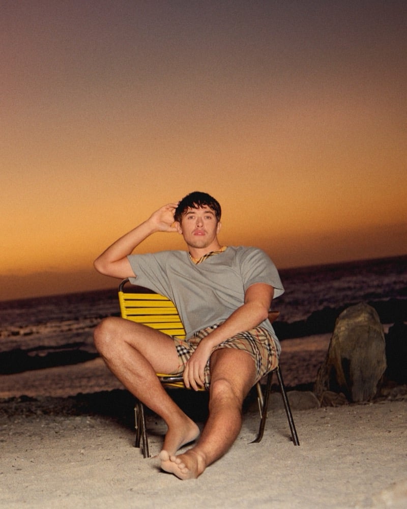 Blyth sits barefoot in a yellow chair on a beach at dusk wearing a gray top and Burberry check shorts against an orange and purple sky for the high summer 2026 campaign