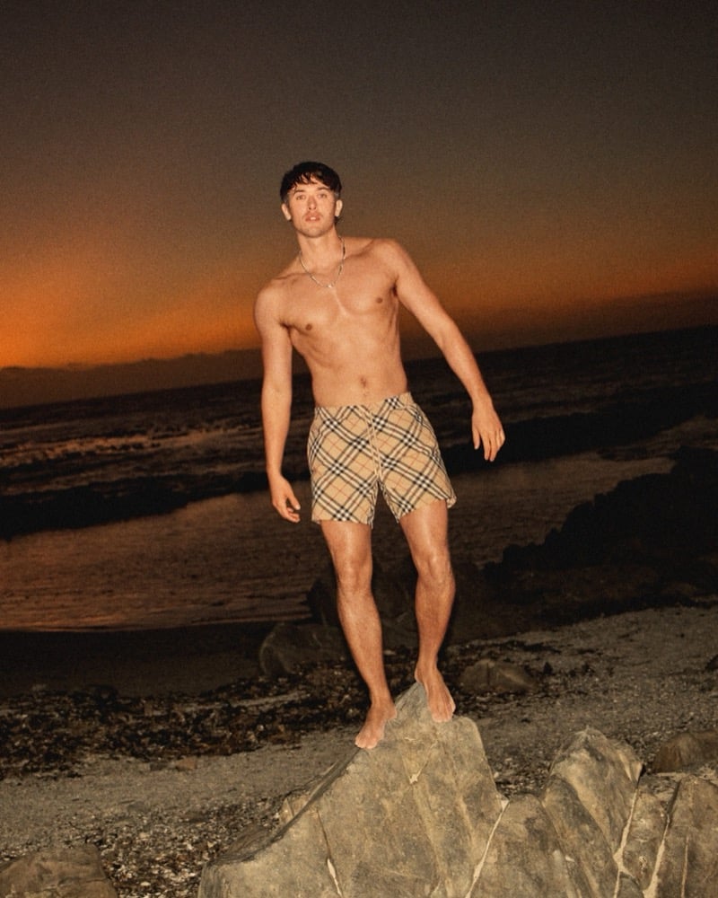 Tom Blyth stands shirtless on rocks at the shoreline wearing Burberry check swim shorts and a chain necklace against an orange sunset sky for the high summer 2026 campaign