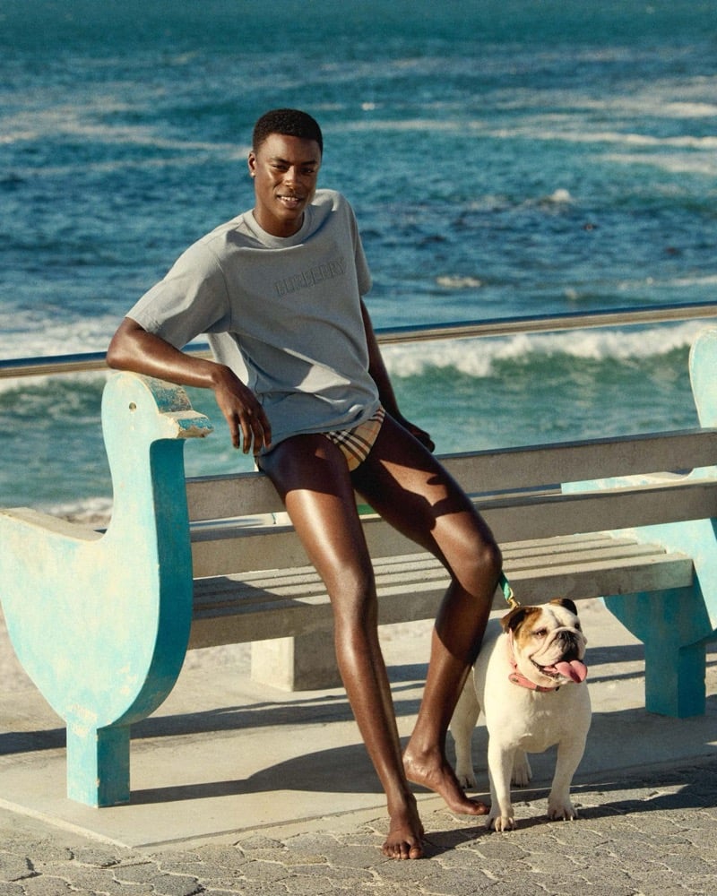N'Doye sits on a pale blue bench near the shoreline in a grey Burberry logo T-shirt and check shorts, with an English bulldog beside him, for the high summer 2026 campaign