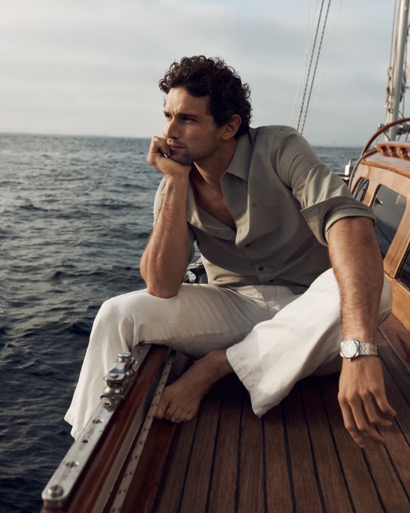 Giacomo sits on the deck of a wooden sailboat in an open green Calvin Klein linen shirt and white linen trousers looking out to sea for the Getaway edit