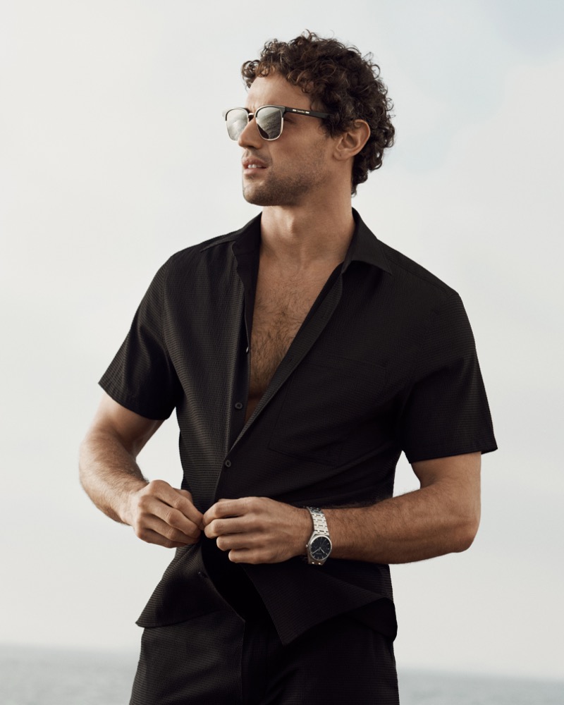 Giacomo buttons a black Calvin Klein seersucker short-sleeve shirt while wearing matching black seersucker trousers and oversized sunglasses for the Getaway edit