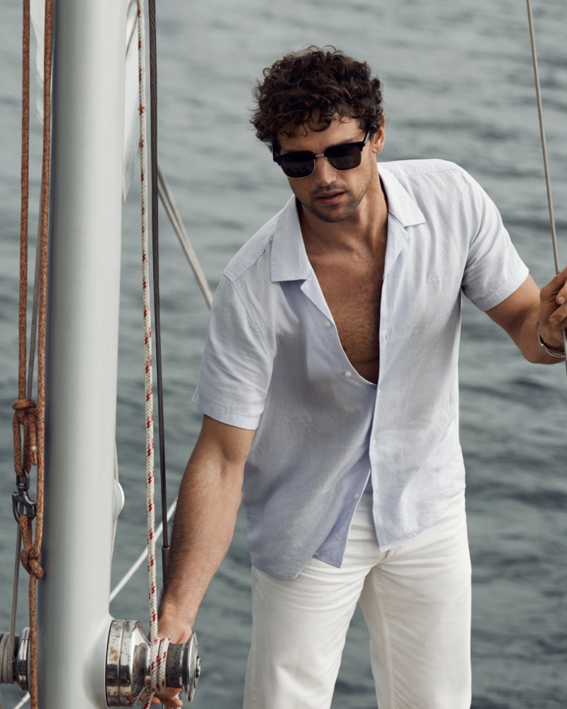 Giacomo grips the rigging of a wooden sailboat in an open light blue Calvin Klein linen camp collar shirt and white trousers for the Getaway edit