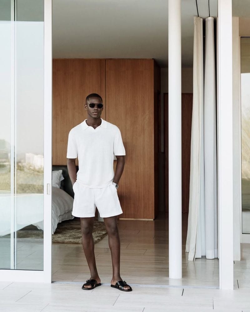 Cheikh wears a white linen polo shirt, white shorts, black sandals, and aviator sunglasses in an open doorway for Calvin Klein spring 2026