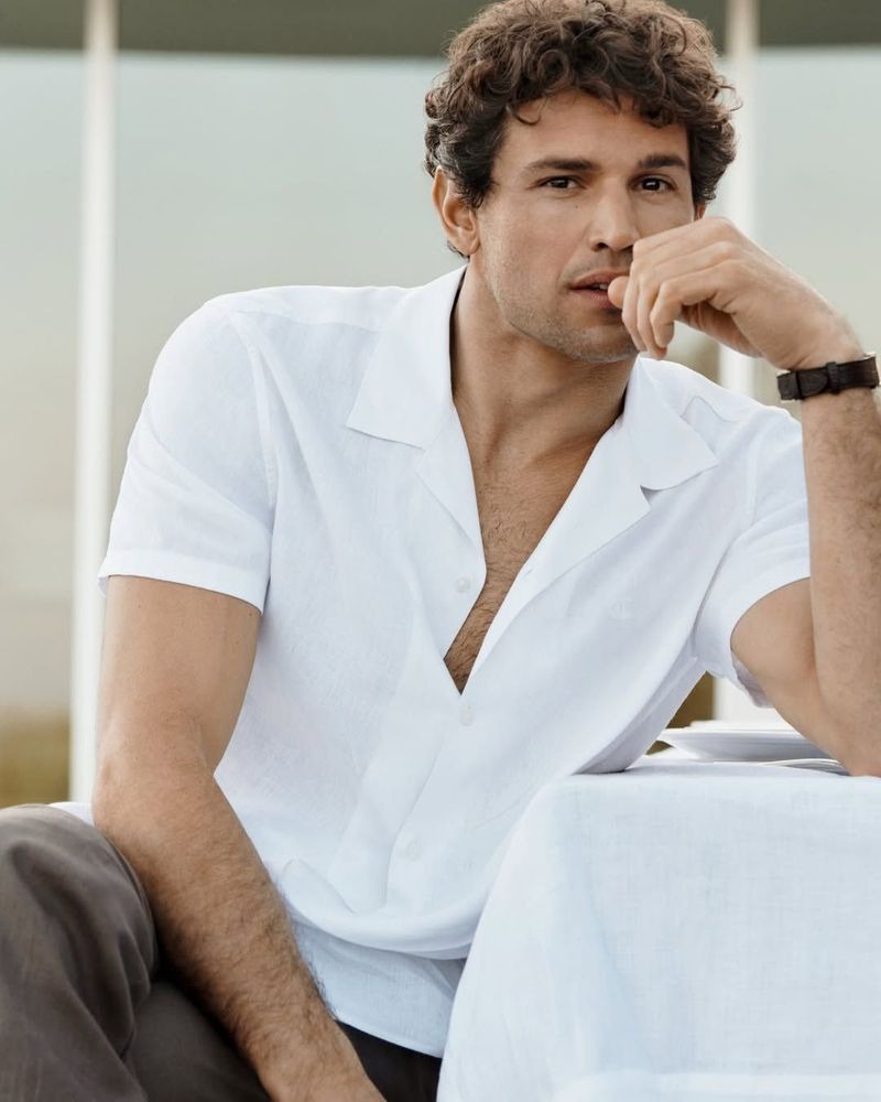 Giacomo sits in a white linen camp-collar short-sleeve shirt left partly open at the chest for Calvin Klein spring 2026