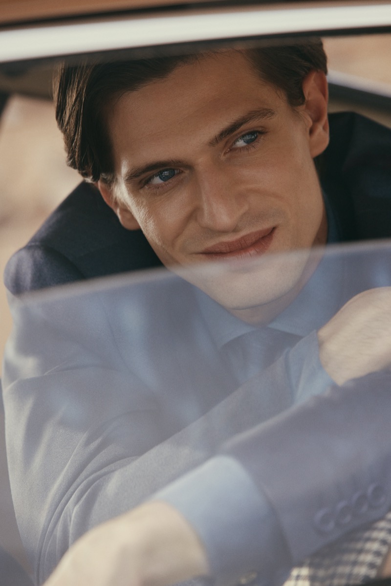 Edoardo smiles through the window of a vintage car in a blue-toned suit and matching shirt, a hint of houndstooth visible below, for Canali's spring-summer 2026 campaign