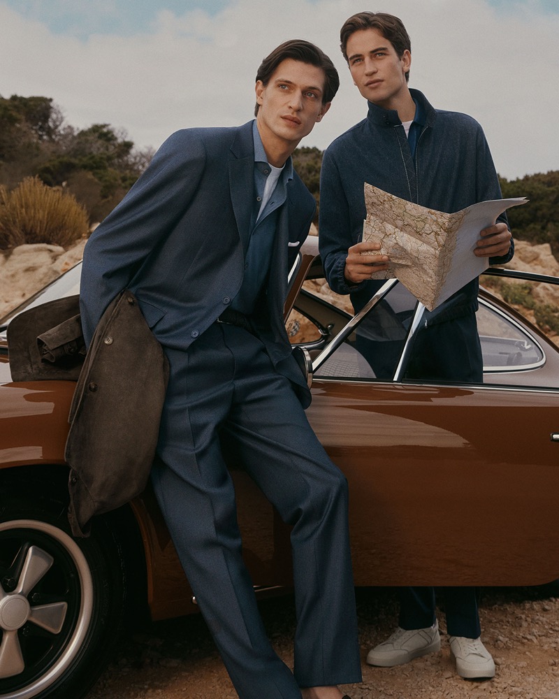 Edoardo leans against a vintage orange grand tourer in a navy blue suit and denim-blue shirt while Luca stands beside him in a navy zip blouson and dark jeans, holding an open map, for Canali's spring-summer 2026 campaign