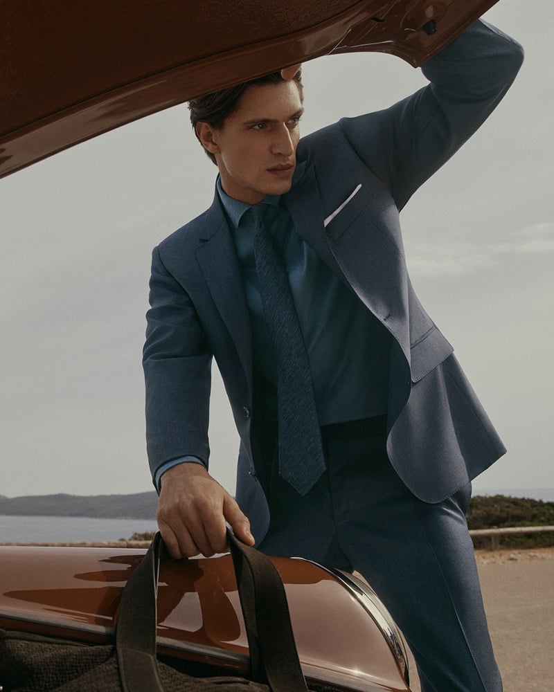 Edoardo stands at the open trunk of a vintage car along a coastal horizon in a mid-blue suit, matching blue shirt, and dark tie for Canali's spring-summer 2026 campaign