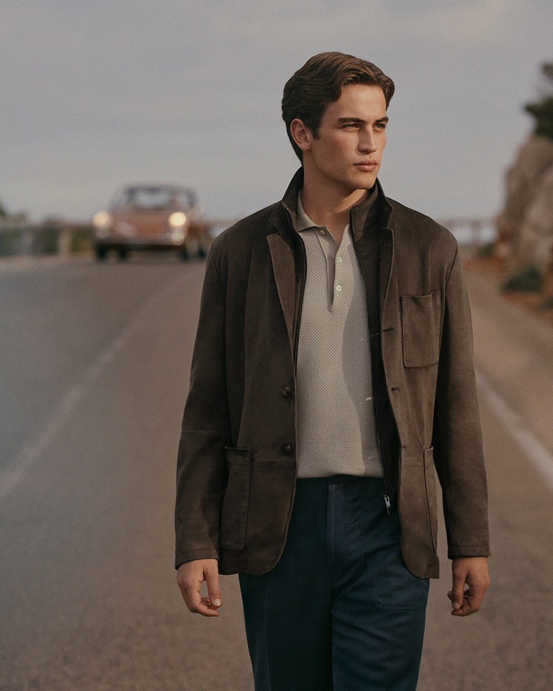 Luca walks on a coastal road in a brown suede blazer over a cream knit polo and navy trousers, a vintage car in the distance behind him, for Canali's spring-summer 2026 campaign