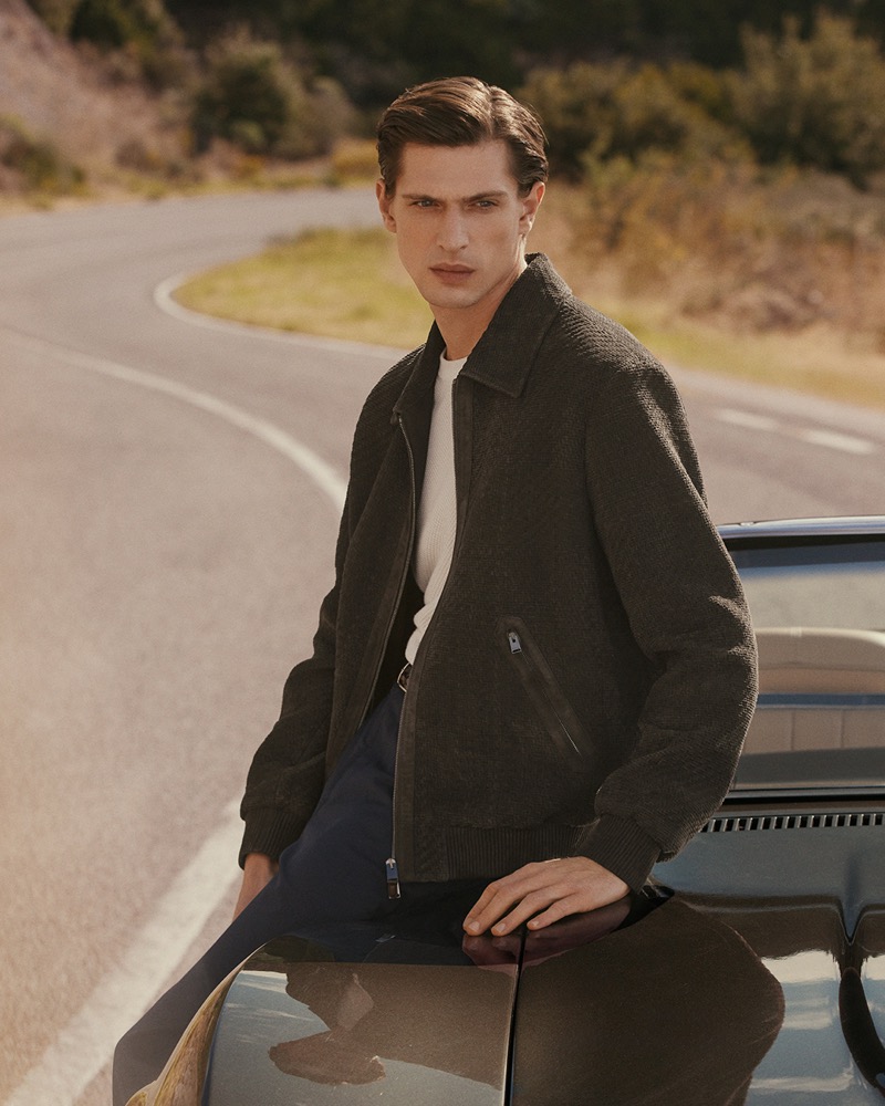 Edoardo sits on a vintage car along an empty mountain road in a charcoal textured zip blouson, white tee, and navy trousers for Canali's spring-summer 2026 campaign