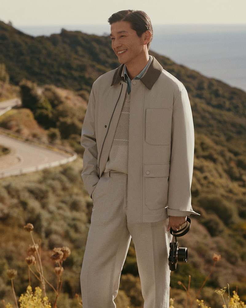 Daisuke stands on a Mediterranean hillside in a stone field jacket over a cream textured knit and matching trousers, holding a camera, with the coast behind him, for Canali's spring-summer 2026 campaign