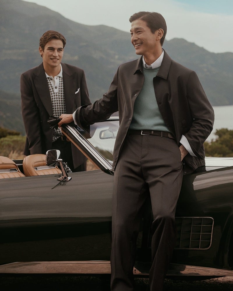 Daisuke stands beside a dark vintage convertible in a dark jacket layered over a teal knit while Luca stands behind the open door in a dark brown blazer over a houndstooth knit polo for Canali's spring-summer 2026 campaign