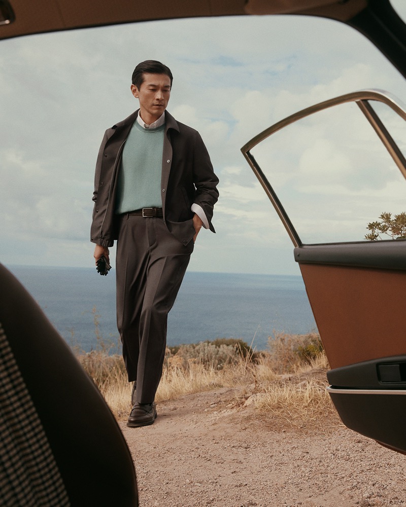 Daisuke walks toward the open door of a vintage convertible above a cliffside sea view in a dark snap blouson over a teal knit, with trousers and driving gloves in hand, for Canali's spring-summer 2026 campaign